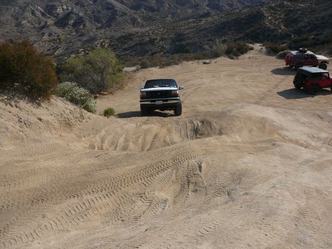 1988 Ford Bronco - A white Ford F-150 navigates a sandy, off-road trail with other vehicles visible in the distance.