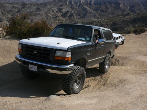 1988 Ford Bronco - A lifted Ford Bronco navigates a dusty off-road trail with another vehicle following in the background.