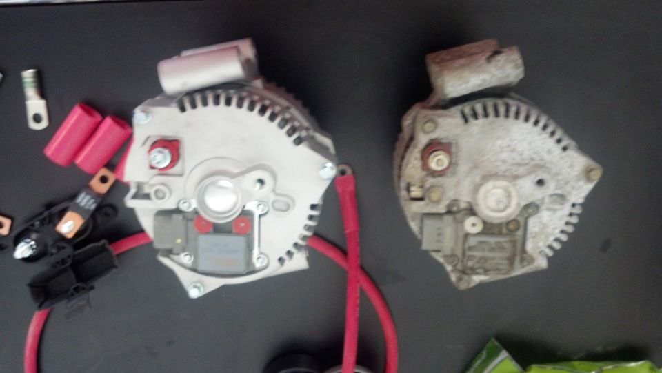 Two automotive alternators, one new and one used, with red cables and electrical connectors on a dark surface