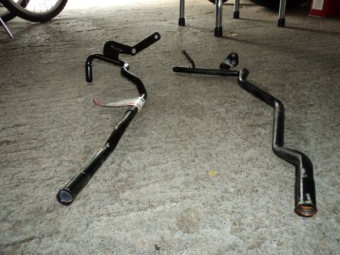 2004 Mercury Grand Marquis - Two black metal pipes, possibly exhaust or coolant lines, lie on a concrete surface. One pipe has a white tag attached.