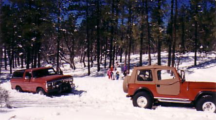 1979 Ford Bronco - Two off-road vehicles are in a snowy forest with people in the background.