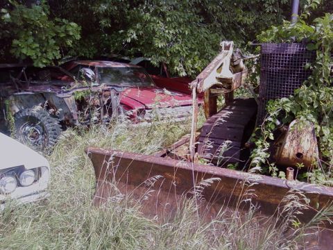 1974 Dodge Challenger - A red car, partially obscured by vegetation, sits in a yard with a bulldozer in the foreground. The Supermotors.net logo is visible in the…