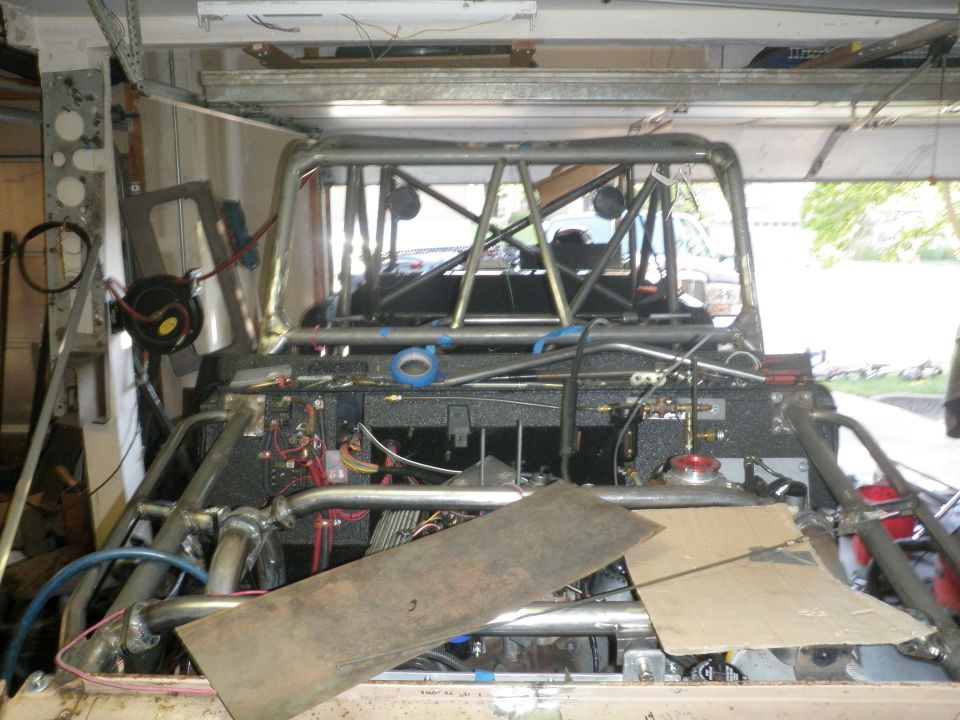 Custom roll cage and engine bay with exposed wiring and plumbing. Cardboard and metal sheets are placed over parts of the engine