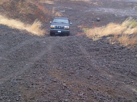 1994 Ford Bronco - A black Ford Bronco navigates a rough, rocky terrain with dry grass on the sides.