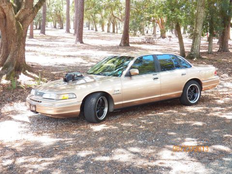 1993 Ford Crown Victoria - A gold 1993 Ford Crown Victoria sedan with a prominent hood scoop and aftermarket wheels sits in a shaded, wooded area.