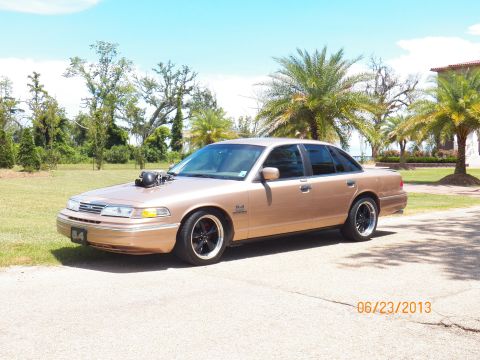 1993 Ford Crown Victoria - A gold sedan with a prominent hood scoop and aftermarket wheels is parked outdoors under a bright sky.
