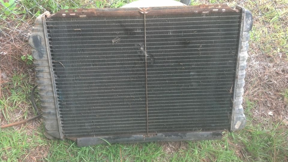 Used, dirty radiator with rusty top mount standing on grass against a chain-link fence