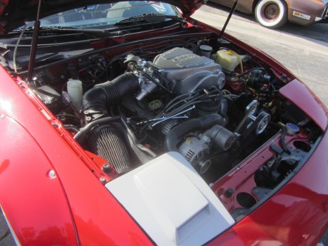 1964 Ford Ranchero - Engine bay of a red car, a custom intake and a prominent silver engine cover, with another vehicle visible in the background.