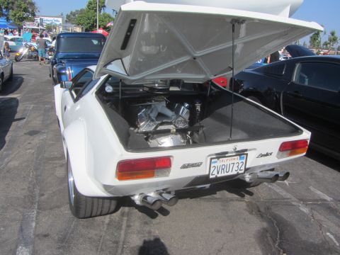 1964 Ford Ranchero - A white sports car with its trunk open, displaying a V8 engine and quad exhaust pipes, parked at an outdoor event.