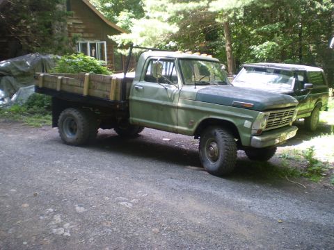 1979 Ford Bronco - A green flatbed truck and a dark SUV are parked on a gravel road in front of a wooden cabin.