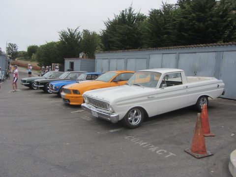 1964 Ford Ranchero - A row of classic cars, including a white 1964 Ford Ranchero, are parked outside a building.