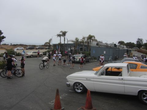 1964 Ford Ranchero - A group of cyclists gather near a white 1964 Ford Ranchero and an orange car in a parking lot.