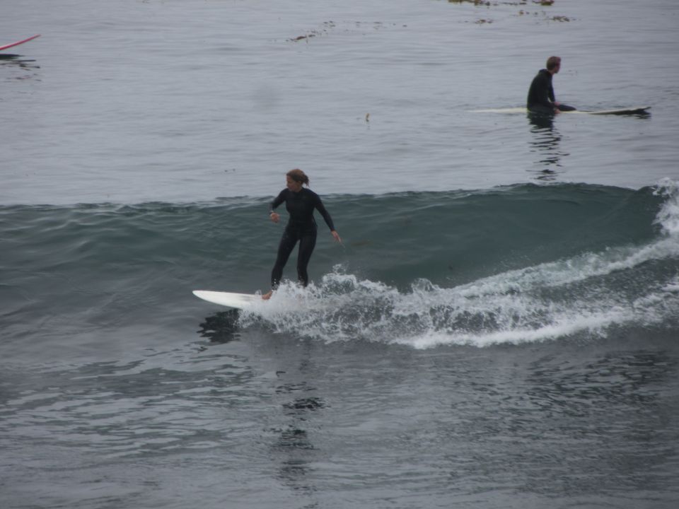 Surfer in a black wetsuit rides a wave on a white surfboard, with another surfer in the background