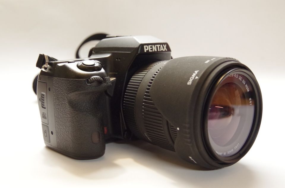 Pentax DSLR camera body with a Sigma zoom lens attached. Text on lens reads 'SIGMA 18-50 DC OS HSM'