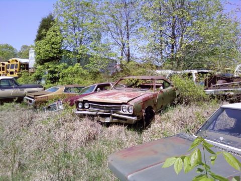 1974 Dodge Challenger - Multiple abandoned vehicles sit in a grassy junkyard under a clear blue sky.