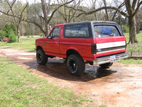 1992 Ford Bronco - A red 1992 Ford Bronco with a black top is shown from a three-quarter front view on a dirt road.