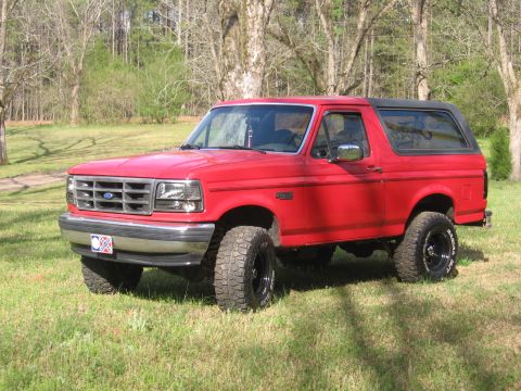 1992 Ford Bronco - Red 1992 Ford Bronco with lifted suspension and aftermarket wheels sits in a grassy, wooded area.