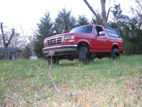 1992 Ford Bronco - A red 1992 Ford Bronco is parked on a grassy incline, with trees and a small building visible in the distance.