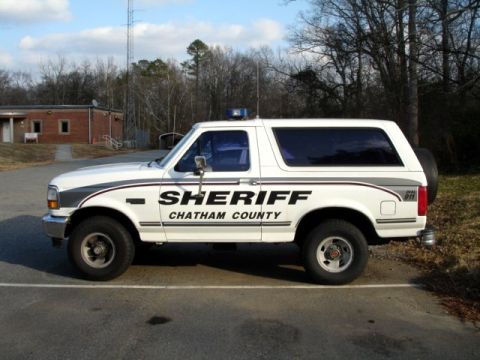 1992 Ford Bronco - A white Ford Bronco SUV, marked as a Sheriff's vehicle for Chatham County, is parked in a lot near a building.