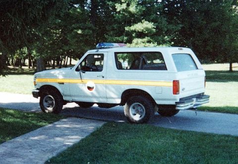 1992 Ford Bronco - A white 1992 Ford Bronco, marked with a yellow stripe and emergency lights, is parked on a paved surface next to a grassy area.
