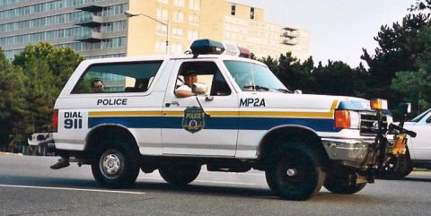 1992 Ford Bronco - A 1992 Ford Bronco police SUV, marked 'POLICE' and 'MP2A', is seen driving on a road with a building in the background.