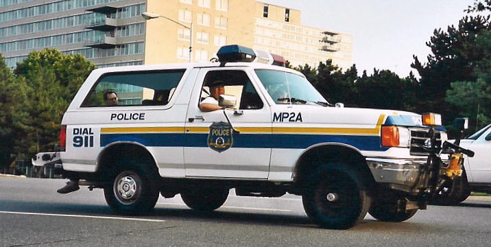 White 1992 Ford Bronco police SUV with blue and yellow stripes, marked 'POLICE' and 'MP2A', driving on a road