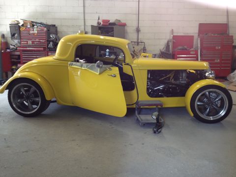 1975 Ford Ranchero - A yellow hot rod coupe sits in a garage with its door open, revealing the interior and engine bay.