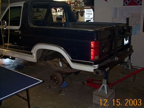 1986 Ford Bronco - Rear view of a dark blue and white Ford Bronco, raised on jack stands for maintenance.