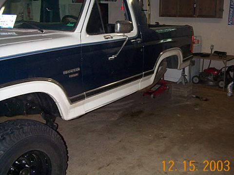 1986 Ford Bronco - A dark blue and white 1986 Ford Bronco is shown from the side, appearing to have a lifted suspension and is supported by a jack.