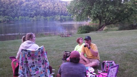 1979 Ford Bronco - A group of people enjoy a riverside gathering with a campfire and drinks.