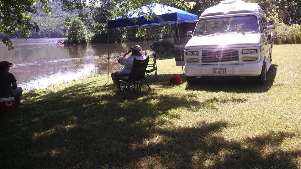 White GMC van parked on grass near a lake, two people sitting in chairs under a blue canopy
