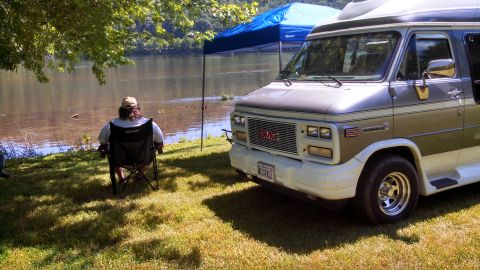 1979 Ford Bronco - A GMC van is parked near a lake while a person relaxes in a chair under a tree.