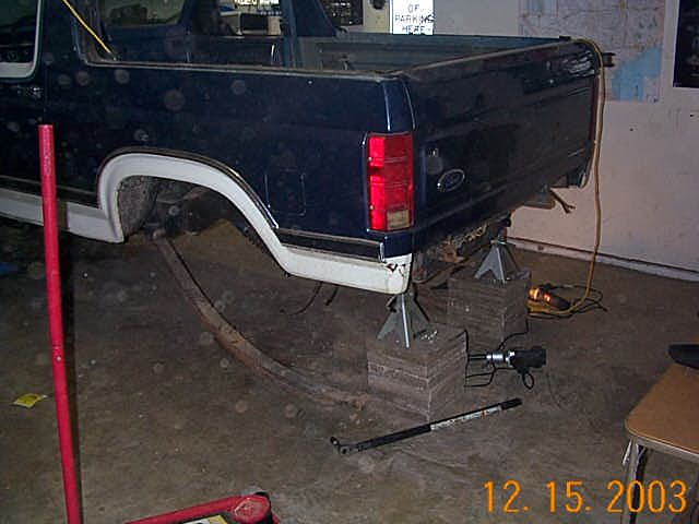 Dark blue Ford truck rear raised on jack stands over wood blocks in a garage.