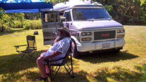 1979 Ford Bronco - A man relaxes in a lawn chair next to a GMC van parked in a grassy area on a sunny day.