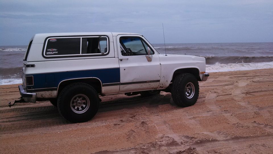 White and blue SUV with a camper shell parked on a sandy beach near the ocean