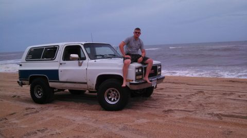 1979 Ford Bronco - A man sits on the hood of a white GMC SUV parked on a sandy beach with the ocean in the background.