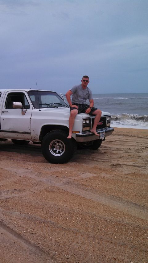 1979 Ford Bronco - Man sits on the hood of a white GMC pickup truck on a beach, with the ocean waves behind.