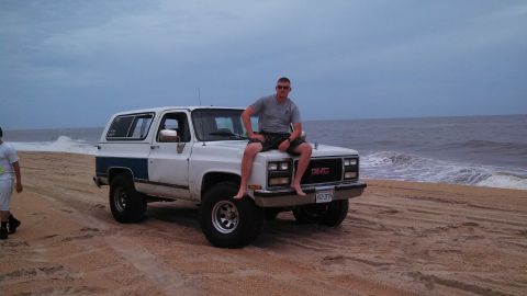 1979 Ford Bronco - A man sits on the hood of a white GMC K5 Blazer parked on a sandy beach with the ocean waves crashing in the background.