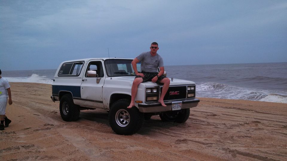 Guy sitting on hood of white GMC K5 Blazer on beach, ocean in background