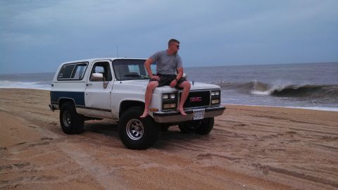 1979 Ford Bronco - A man sits on the hood of a white GMC SUV on a beach with waves in the background.