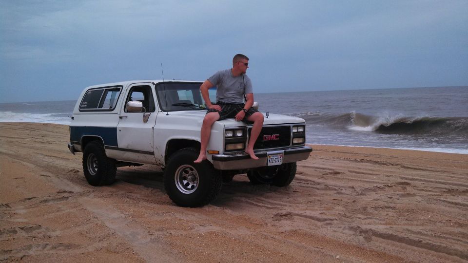 White GMC SUV with blue side trim parked on a sandy beach, man sitting on the hood