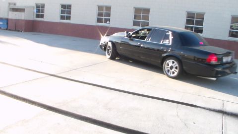 2003 Ford Crown Victoria - black four-door sedan on aftermarket wheels sits parked in a sunlit concrete lot beside a tan building.