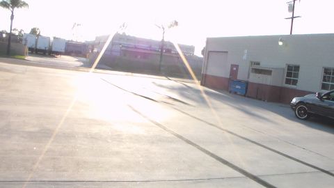 2003 Ford Crown Victoria - dark car sits parked at the edge of a sunlit industrial concrete lot, with strong sun glare across the frame.