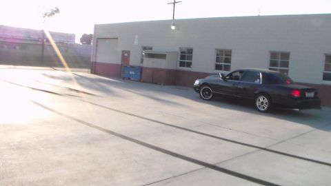 2003 Ford Crown Victoria - black sedan sits parked beside a long industrial building in a concrete lot under a low sun.