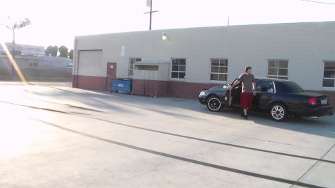 2003 Ford Crown Victoria - man in a t-shirt and red shorts stands beside a black sedan parked in a concrete lot outside an industrial building.