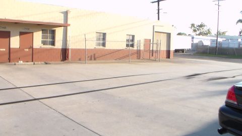 2003 Ford Crown Victoria - open concrete lot in front of a tan industrial warehouse, with a chain-link gate and a parked car edge visible at lower right.