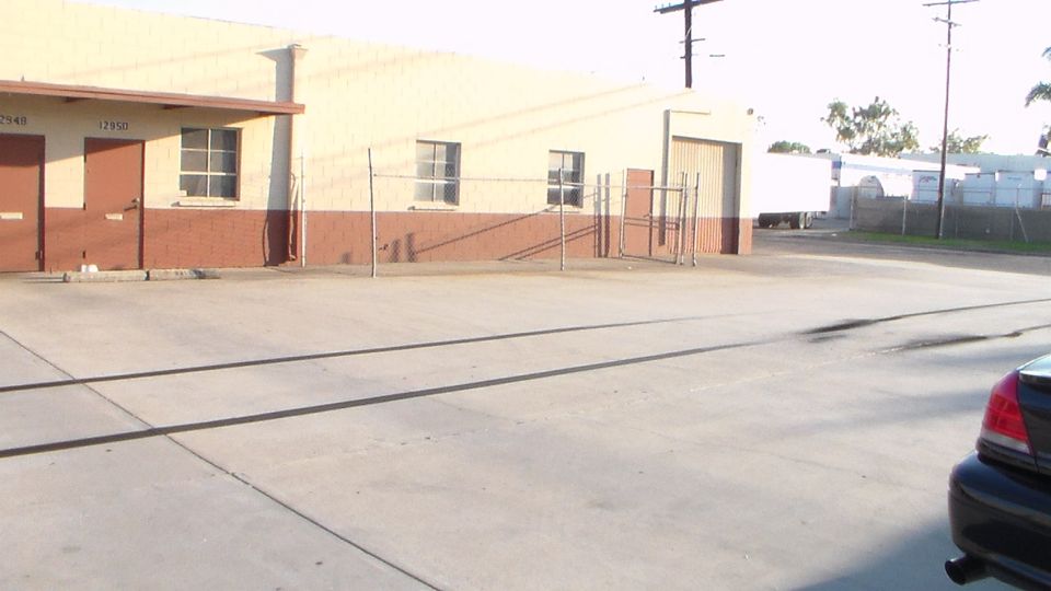 Empty paved industrial yard beside a tan and brick warehouse with chain-link fencing