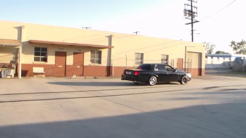 2003 Ford Crown Victoria - black sedan with alloy wheels sits in a wide concrete yard before a long tan industrial warehouse.