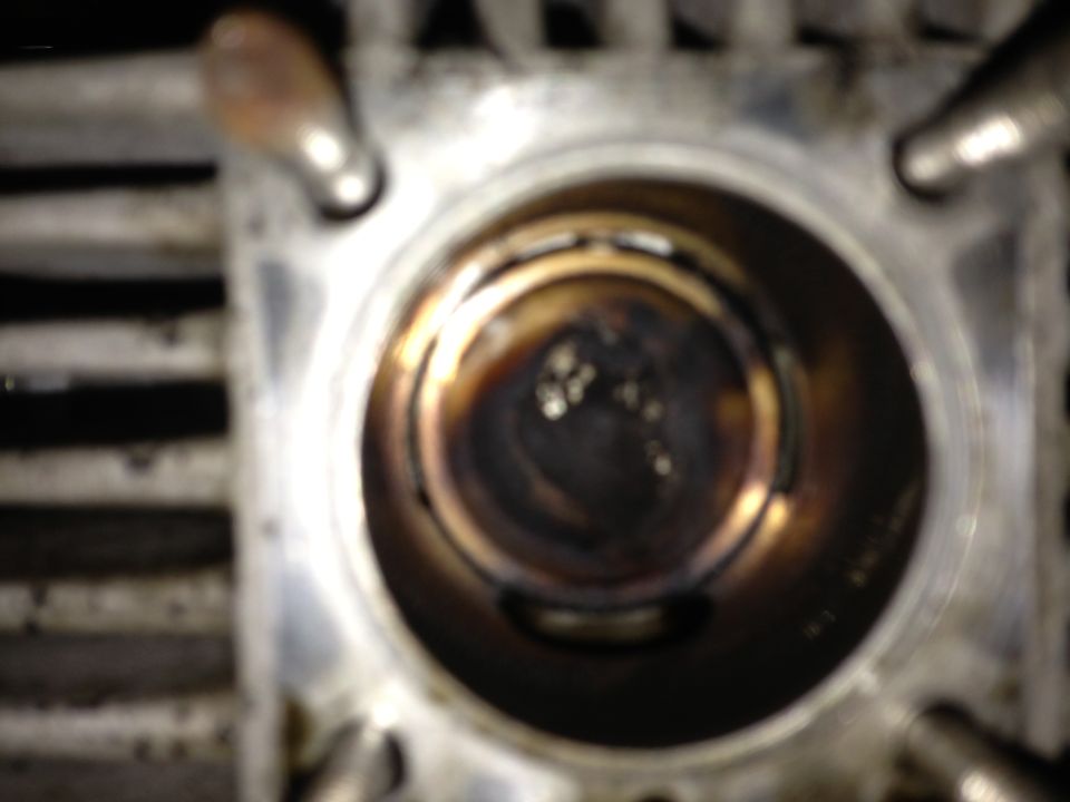 Close-up of an engine cylinder bore with a piston visible inside. The cylinder walls show carbon buildup
