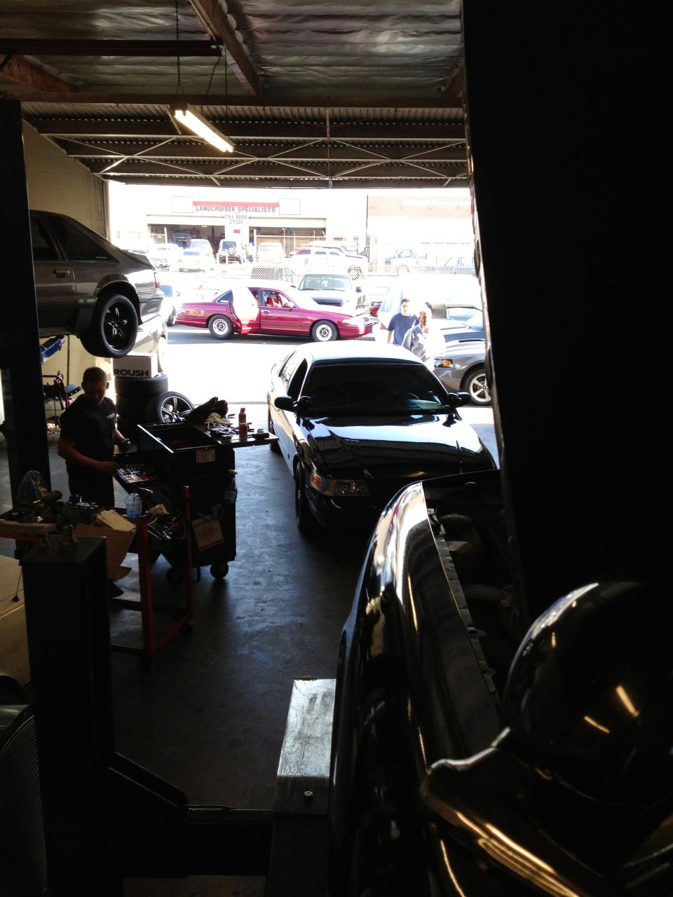 Black sedan on a lift in a garage, with a red car and other vehicles visible outside